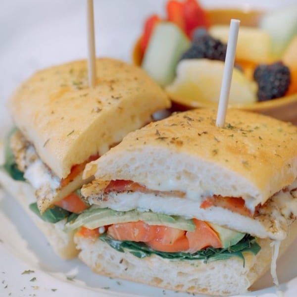 A grilled chicken sandwich with avocado, tomato, and lettuce, served with a side of fruit.