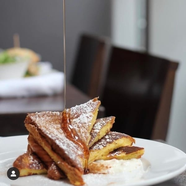 Maple syrup pouring over a stack of powdered sugar-dusted French toast triangles on a white plate.