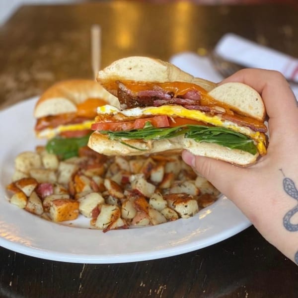 A hand holding a breakfast bagel sandwich with egg, bacon, tomato, and spinach, served with potatoes.