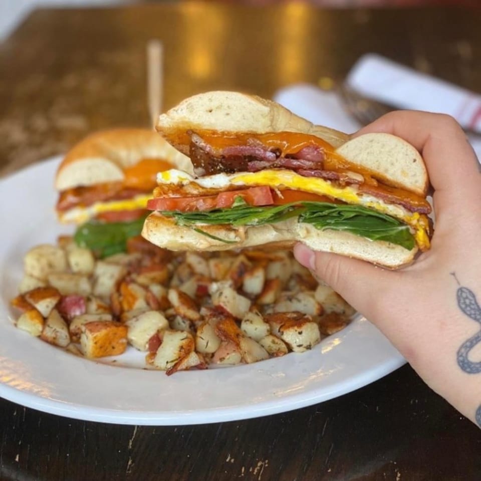A hand holding a breakfast bagel sandwich with egg, bacon, tomato, and spinach, served with potatoes.