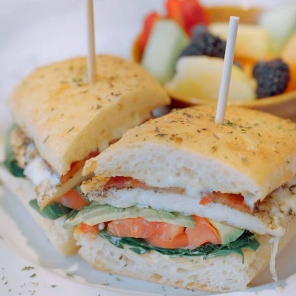 Kanela Breakfast Club's chicken sandwich with avocado, tomato, and lettuce, served with fruit.
