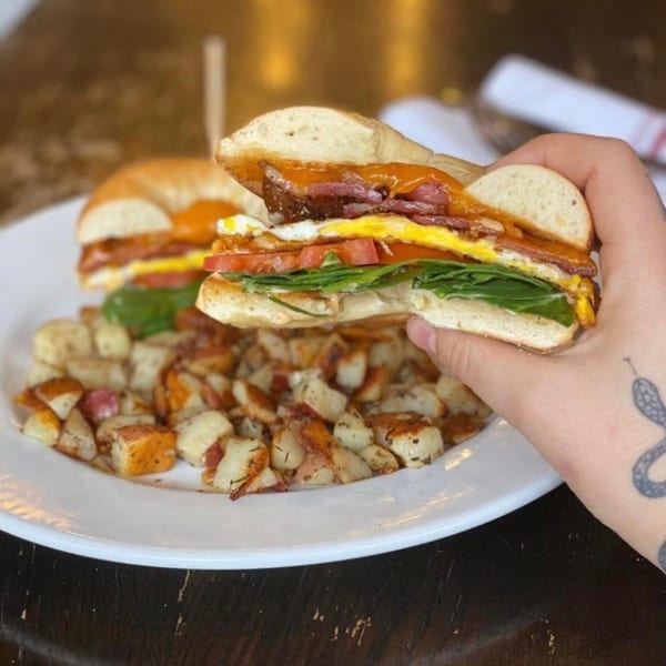 Hand holding a breakfast sandwich with egg, bacon, cheese, tomato, and spinach, served with potatoes.