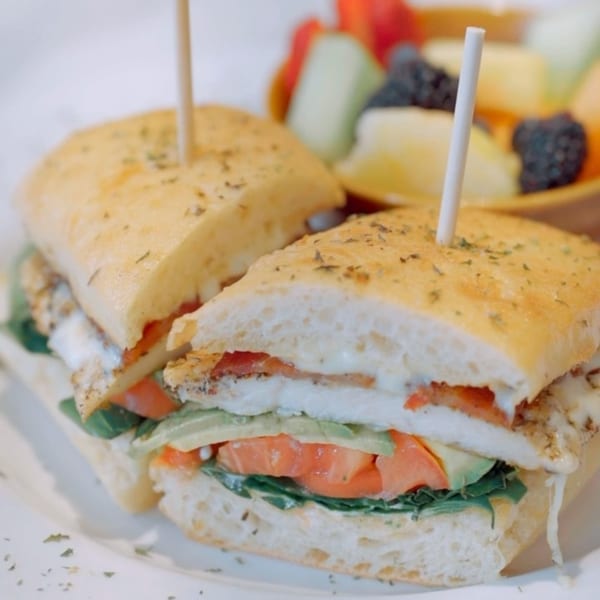 A Kanela Breakfast Club sandwich with egg, avocado, tomato, and greens, served with fruit.